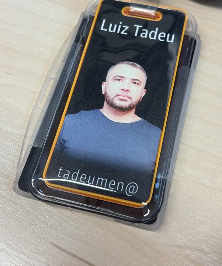An ID badge in a clear plastic holder, lying face-up on a pale wood desk and filling most of the frame: a black card with a yellow border, the name Luiz Tadeu across the top, a portrait photograph below it, and the username tadeumen@ along the bottom.