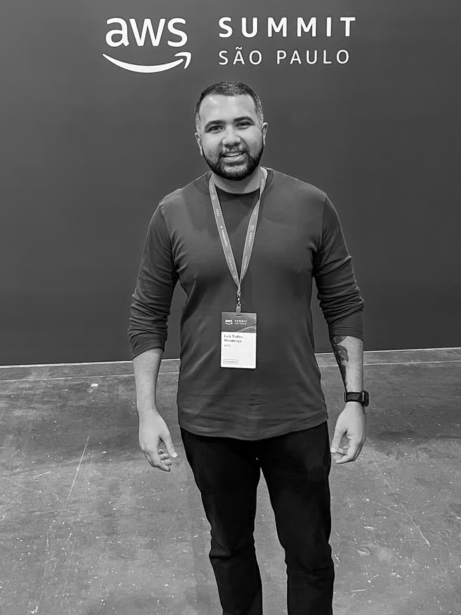 A man standing in front of a plain wall reading "aws Summit São Paulo", a lanyard and badge around his neck.