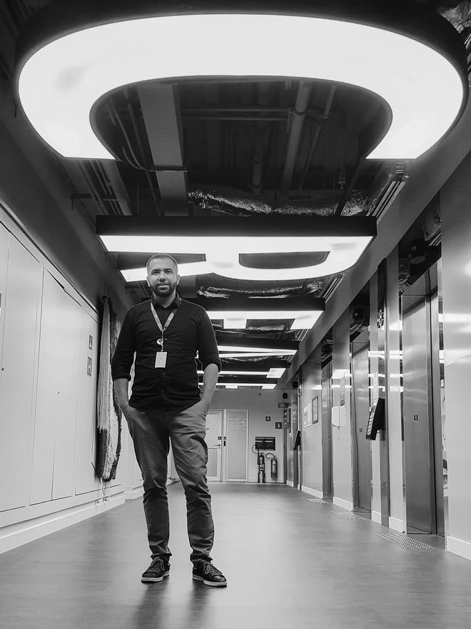 A man standing in the middle of a long, empty office corridor, circular light fittings overhead and the ceiling opened up to show ducts and pipework; lift doors along the right.