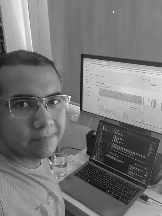 A man in glasses in the foreground, turned to the side, with a desk behind him: an external monitor showing a monitoring dashboard of charts, and an open laptop showing a dark-theme code editor.