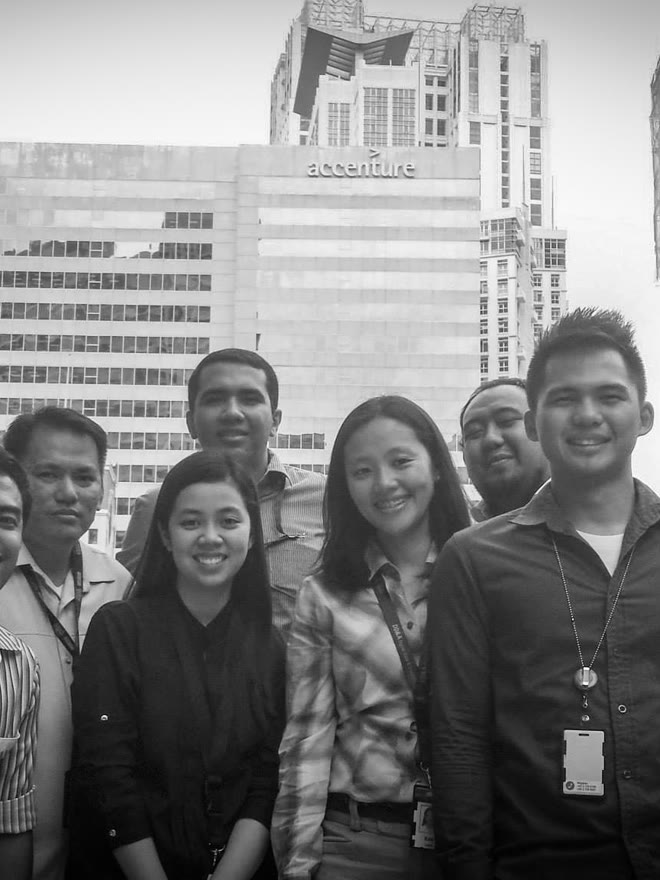 A group of people in front of an office building reading "accenture": five standing across the front — the one at the left edge only half in frame — and two more behind them, smiling at the camera; some with badges on lanyards around their necks and tall towers closing the background.