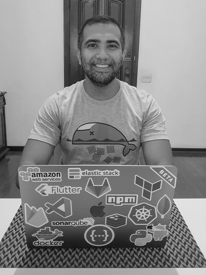 A smiling man behind the open lid of a laptop covered in tool stickers — Amazon Web Services, Elastic Stack, Terraform, Flutter, npm, VS Code, SonarQube, Docker, Kubernetes, MongoDB, Redis, Python, Android — wearing a t-shirt of a whale carrying containers.